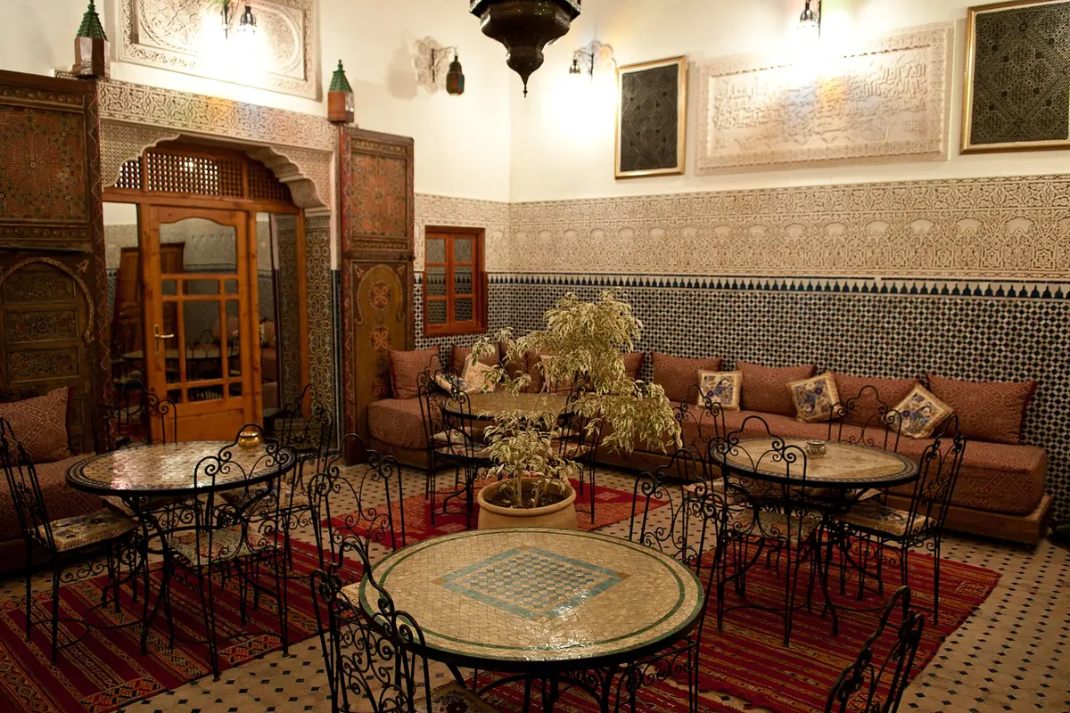 A tiled room in a Fes riad: a band of tile on the lower walls, carved plaster above it, a carved cedar door and small round tables.