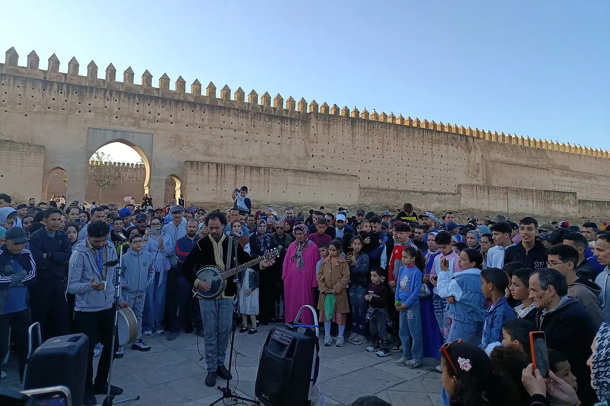 A street musician with a stringed instrument plays in Bab Boujloud Square, Fes, with a crowd gathered in front of the long city wall.