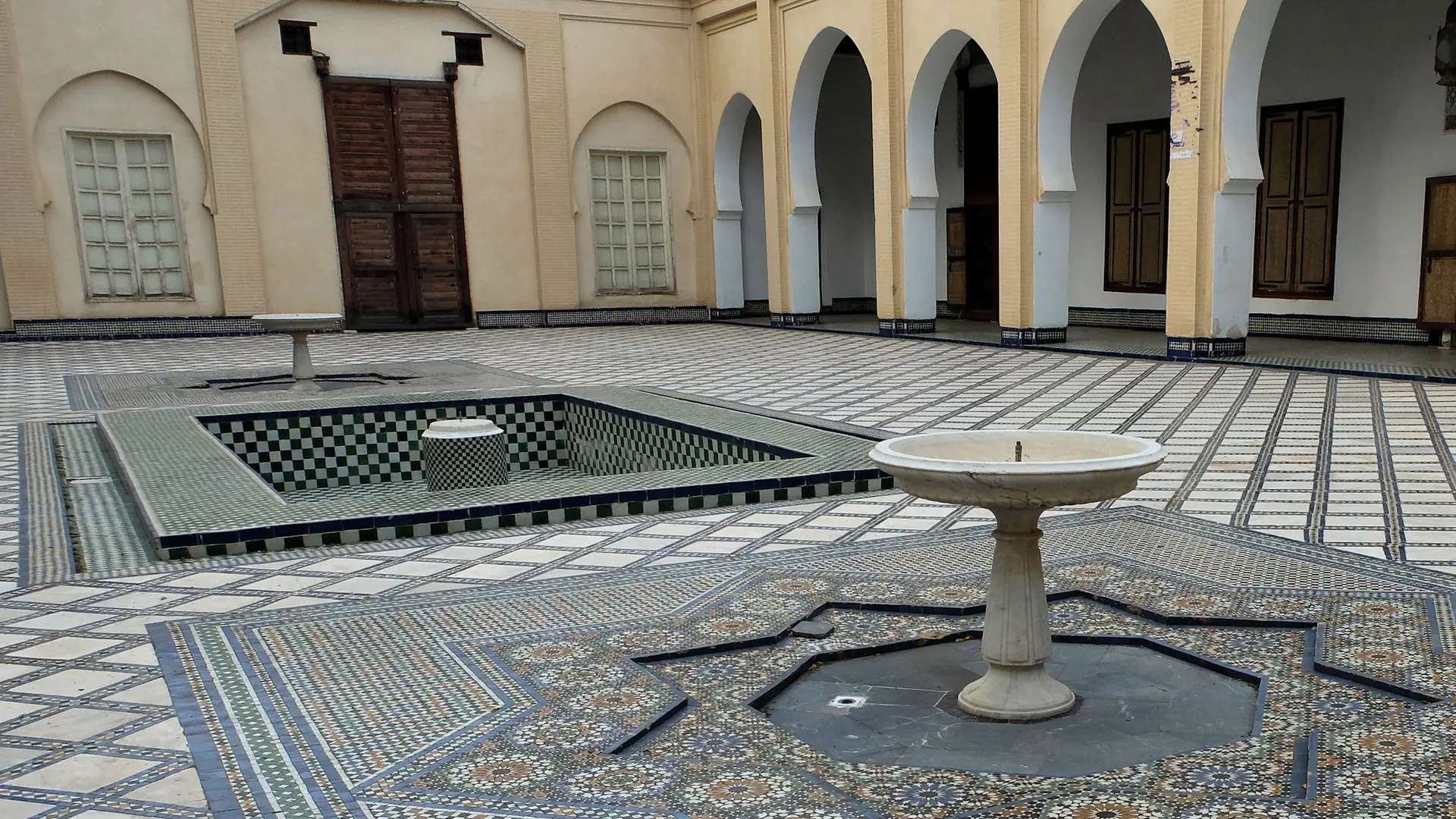 A courtyard in a historic Fes palace, paved in zellij tile with a stone fountain at its centre, under a white arcade of horseshoe arches.